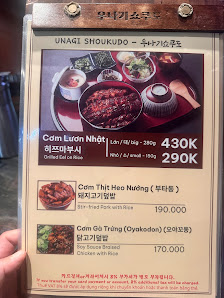 A menu board for 'UNAGI SHOUKUDO' in Thao Dien, Saigon, featuring Vietnamese dishes with prices in K (thousands of VND). The board displays three main