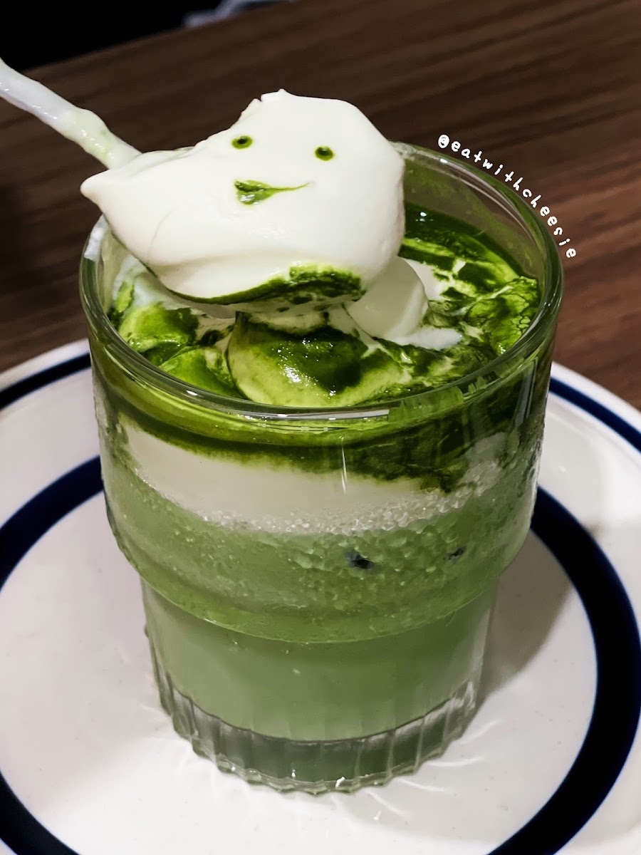 A layered green tea dessert topped with a dollop of cream and sliced cucumbers in a glass cup.