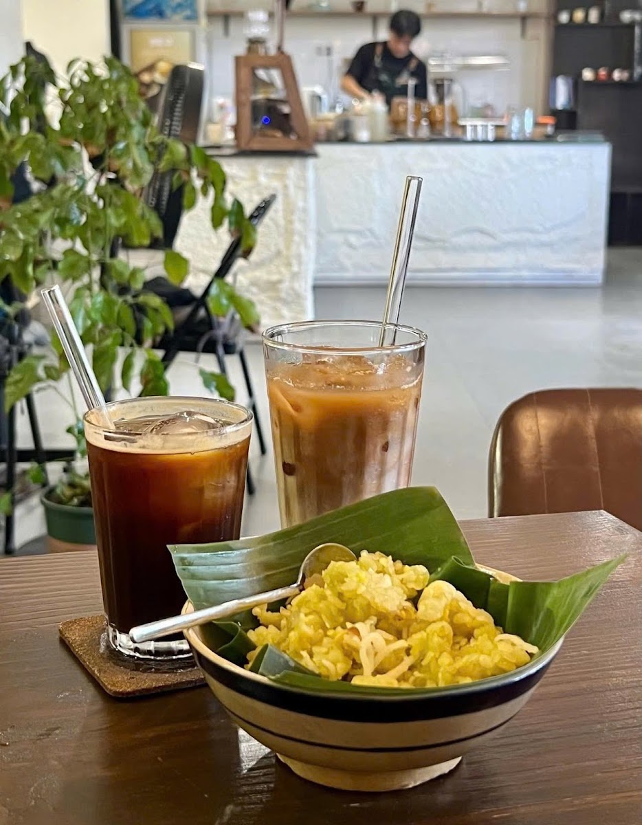 The image shows a table in an indoor cafe setting with two glasses of cold coffee drinks and a bowl containing what appears to be yellow noodles or ri