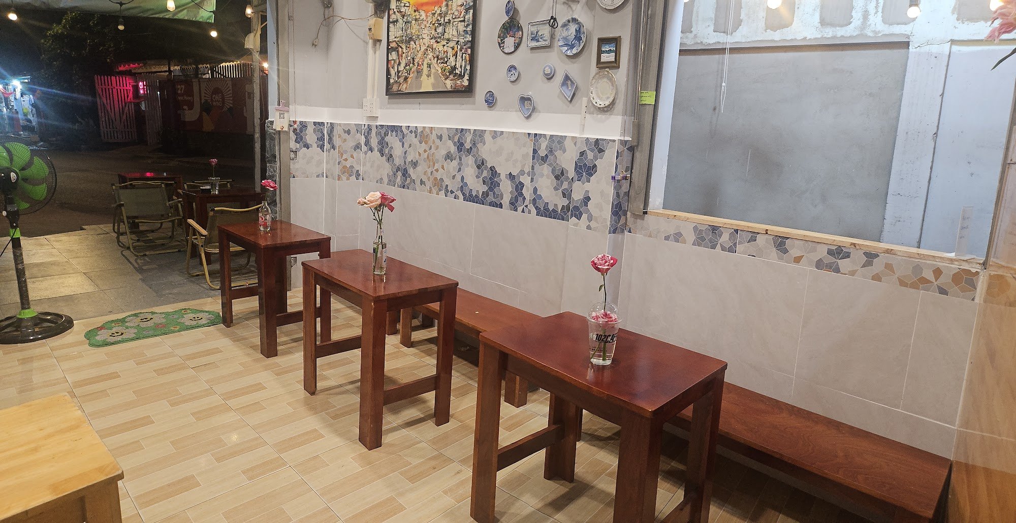 The image shows an interior view of a casual dining establishment at night. The space features wooden tables and chairs with small flower arrangements