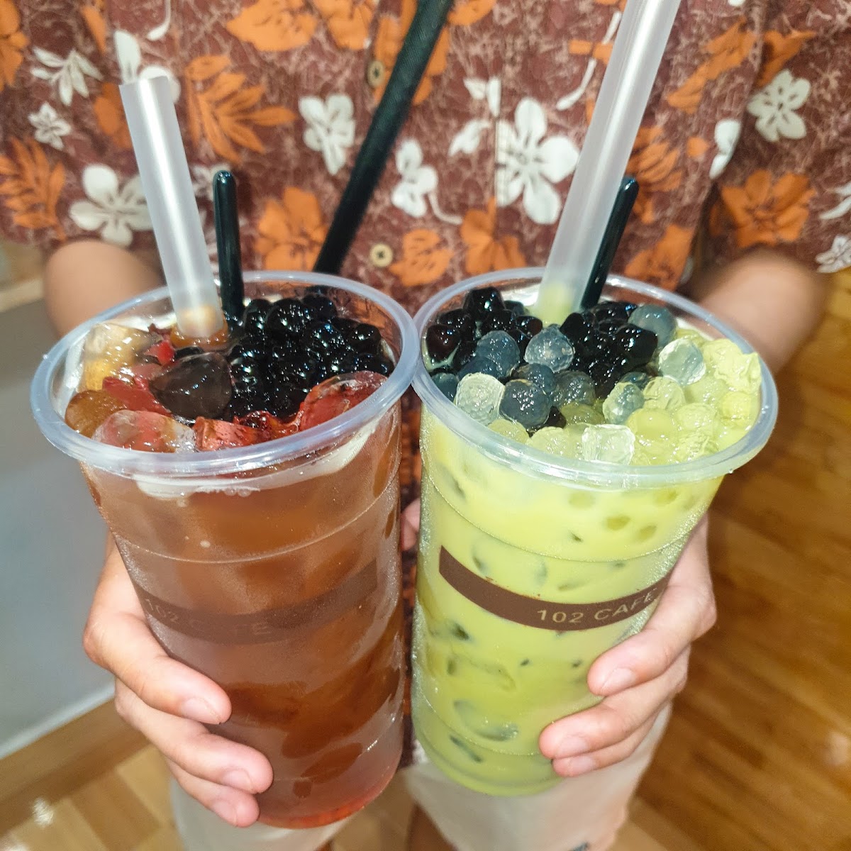 A person is holding two beverages in clear plastic cups with straws and tapioca pearls visible at the bottom of each cup.