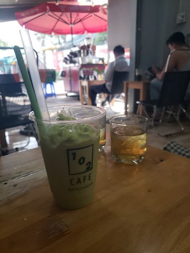 A beverage from '102 Cafe' is placed on a wooden table in an indoor setting with patrons seated at tables and red umbrellas visible, suggesting it mig
