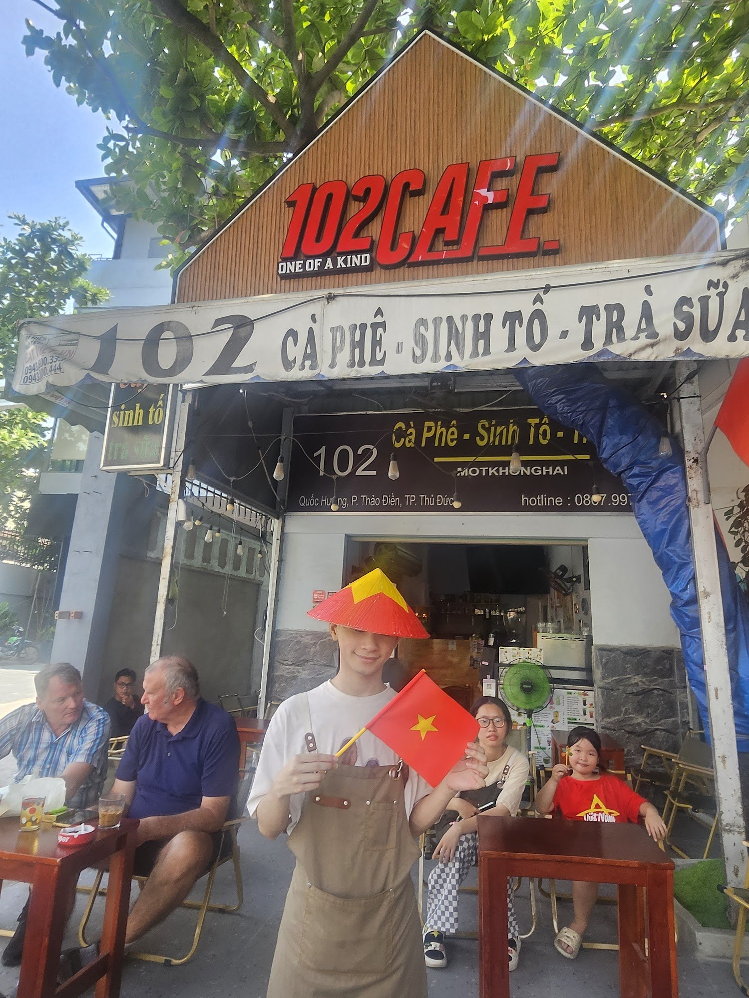 The image shows an outdoor seating area of a cafe named '102 Cafe' with patrons sitting at tables under trees, and one individual standing in the fore