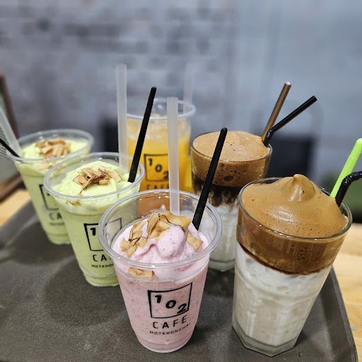 A collection of various beverages served in clear plastic cups on a tray at '102 Cafe' with straws and toppings such as nuts or whipped cream.