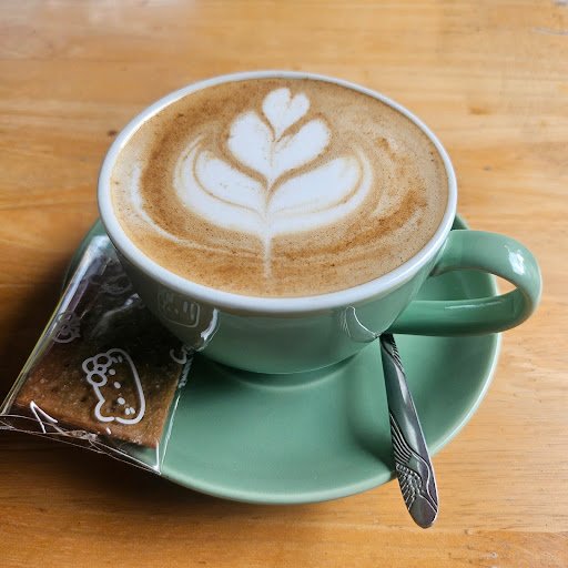 A latte art in a green cup on a saucer accompanied by a cookie and a spoon with intricate designs.
