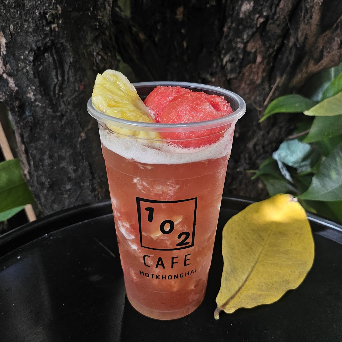 A refreshing drink from '102 Cafe Motkhonghail' served in a clear plastic cup with the cafe's logo on it, garnished with pineapple and watermelon piec