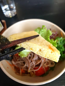 A close-up of a bowl containing noodles and vegetables topped with tofu being lifted by chopsticks.