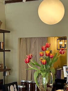 The image shows a cozy interior setting featuring a bouquet of red and yellow tulips in a vase on the dining table, with jars possibly containing pres