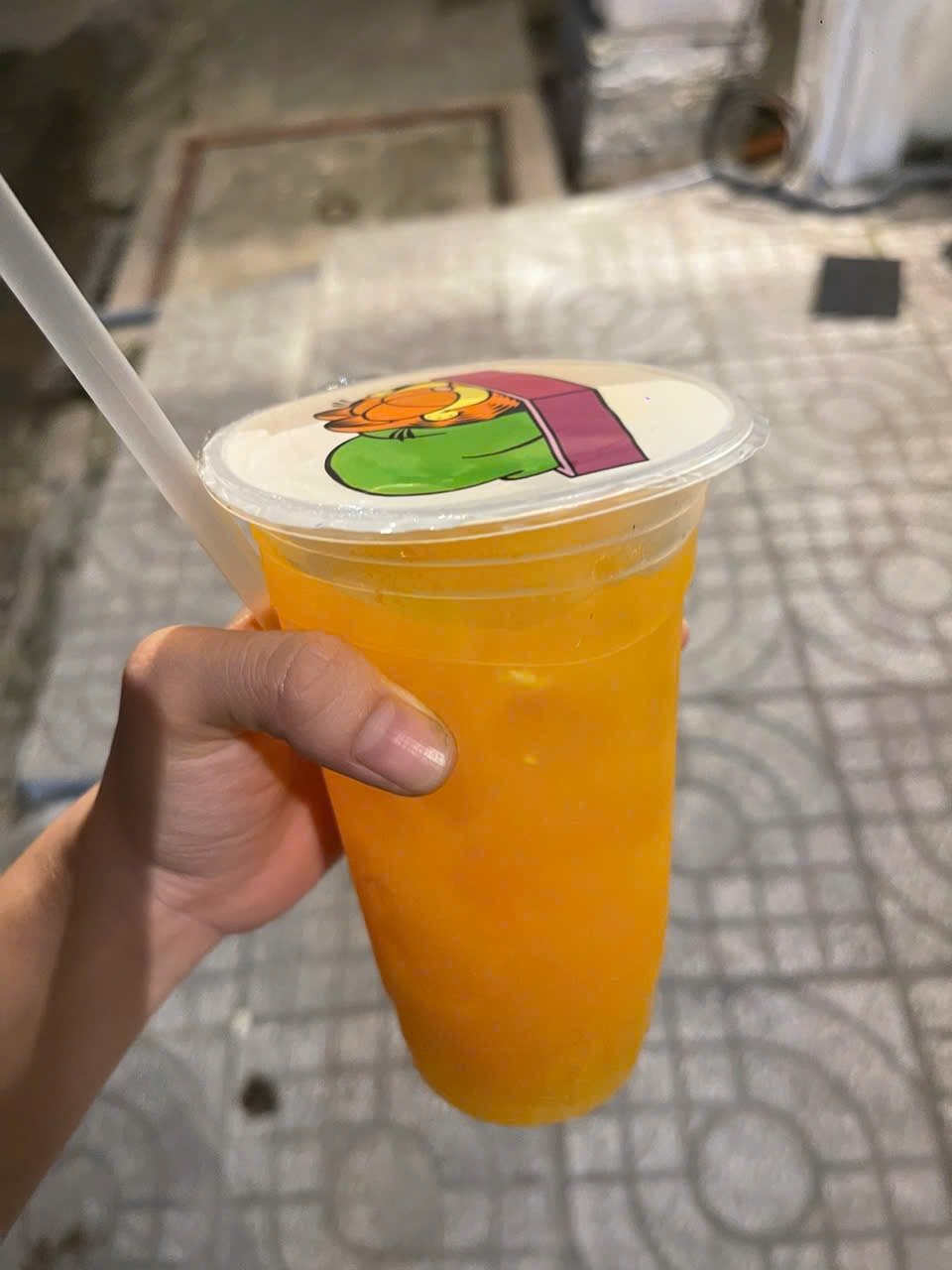 A hand is holding a plastic cup filled with an orange-colored beverage that has a white lid featuring cartoon characters in green and purple, along wi