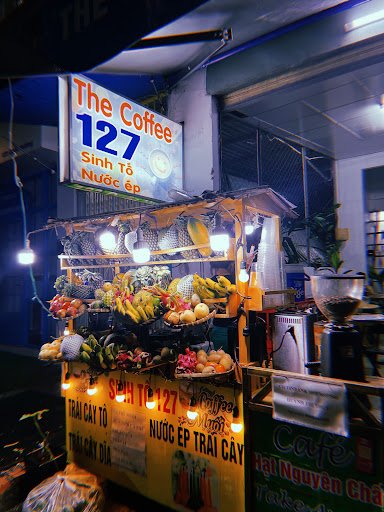 A brightly lit coffee cart named 'The Coffee 127' is situated on a street at night with various fruits and beverages displayed for sale.