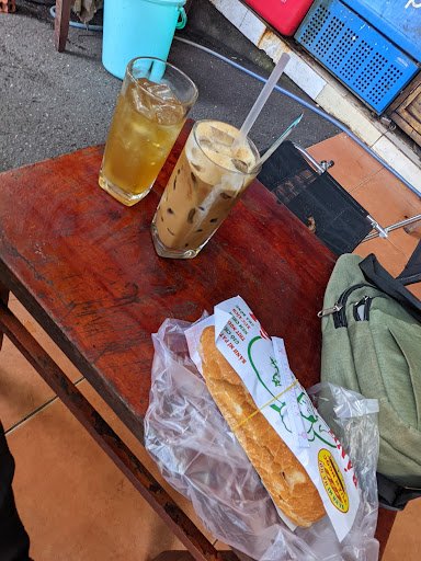 The image shows a wooden table on which two glasses of cold beverages and a wrapped sandwich are placed. The setting appears to be outdoors, possibly