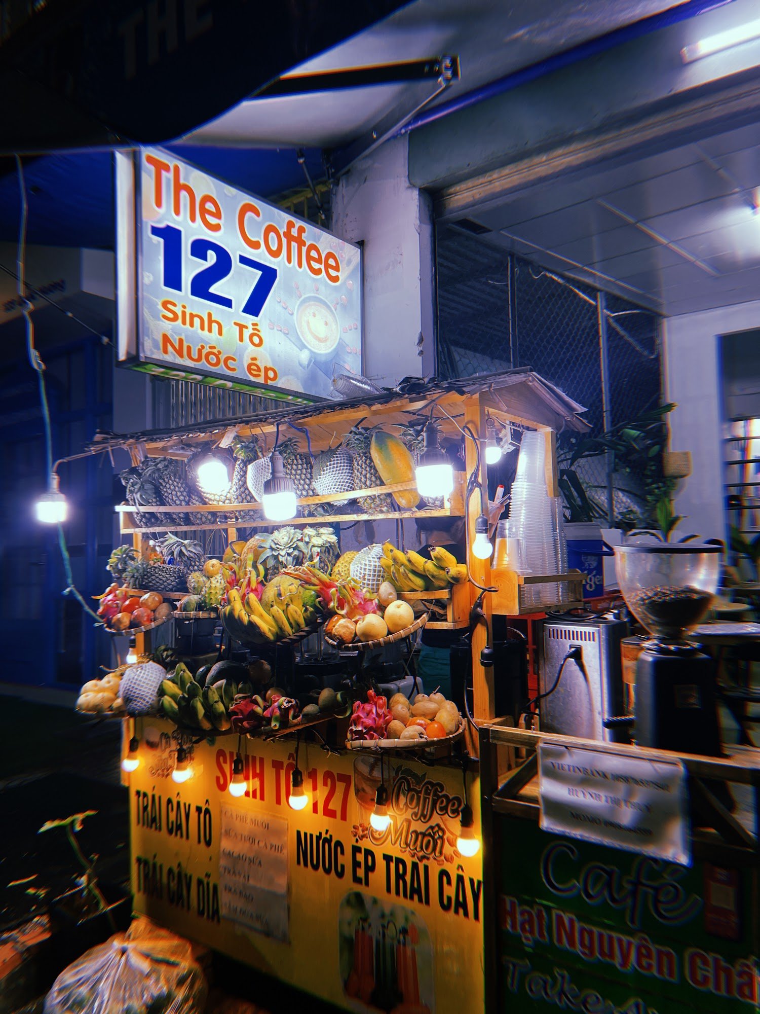 The image shows a brightly lit food stall named 'The Coffee 127' in Thao Dien, Saigon. The stall is adorned with various fruits and has an array of li
