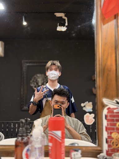 In the image, a person is taking a selfie in front of a mirror in a restaurant interior. Another person stands behind, wearing a mask and apron. The b