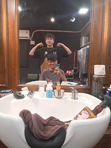 A bathroom interior featuring a white sink with various toiletries (bottles, soap, towel), a wooden-framed mirror reflecting two people taking a selfi