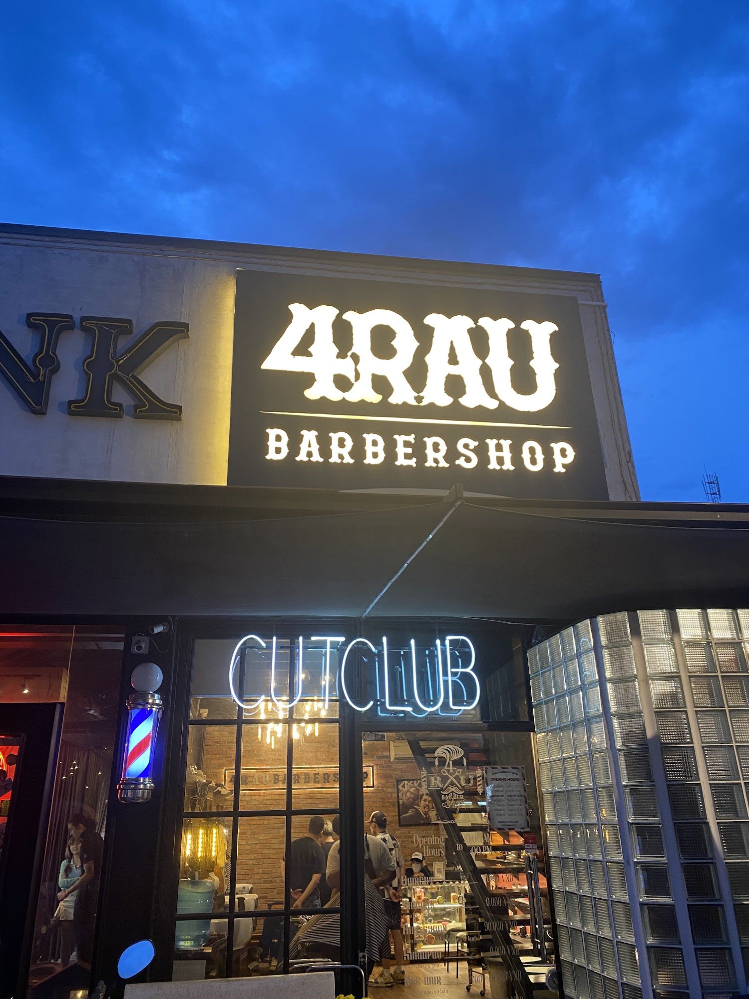 The image depicts the exterior of a barbershop named '4RAU BARBERSHOP' at dusk, with a prominent illuminated sign. Below the main sign, a neon 'CUTCLU