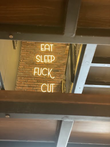 The image shows a neon sign with the text 'EAT SLEEP FUCK CUT' mounted on a brick wall, viewed through a wooden and metal structural frame. The settin