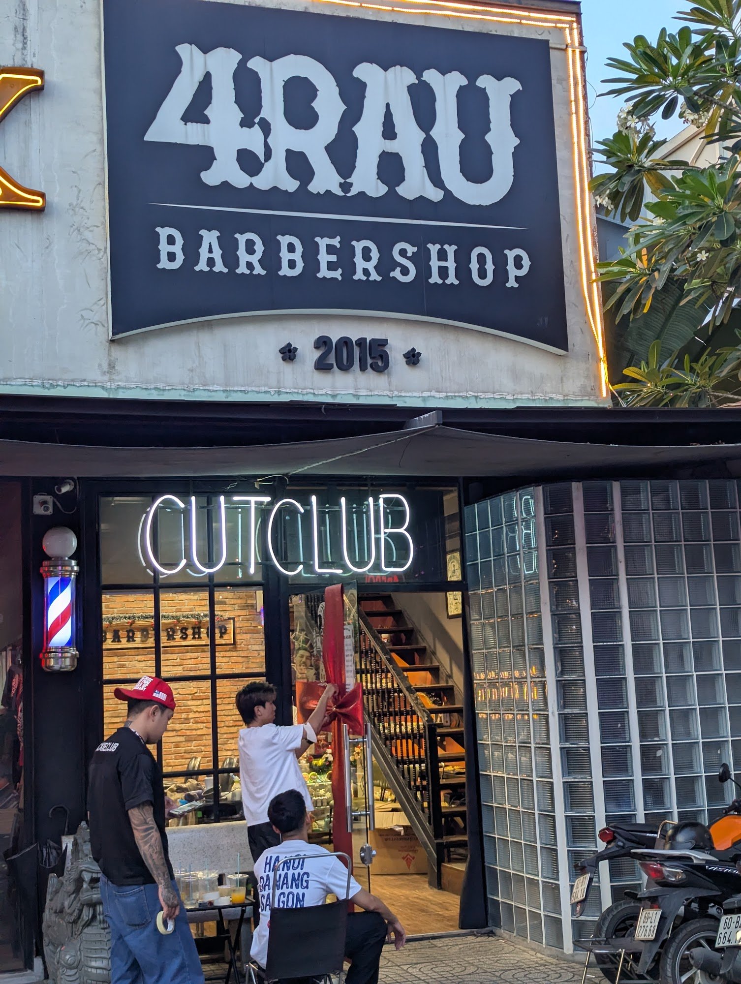 The image depicts the exterior of a barbershop named '4RAU BARBERSHOP' established in 2015, featuring a prominent neon sign 'CUTCLUB' above the entran
