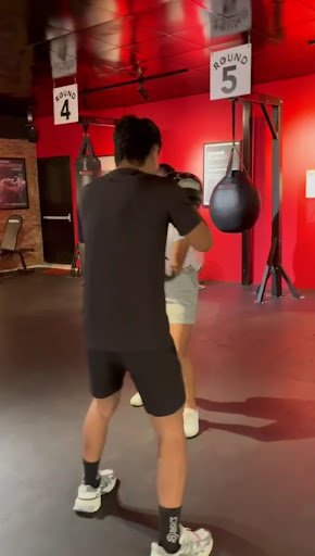 The image shows an indoor gym setting where a person is engaged in boxing training under the supervision of another individual who appears to be provi