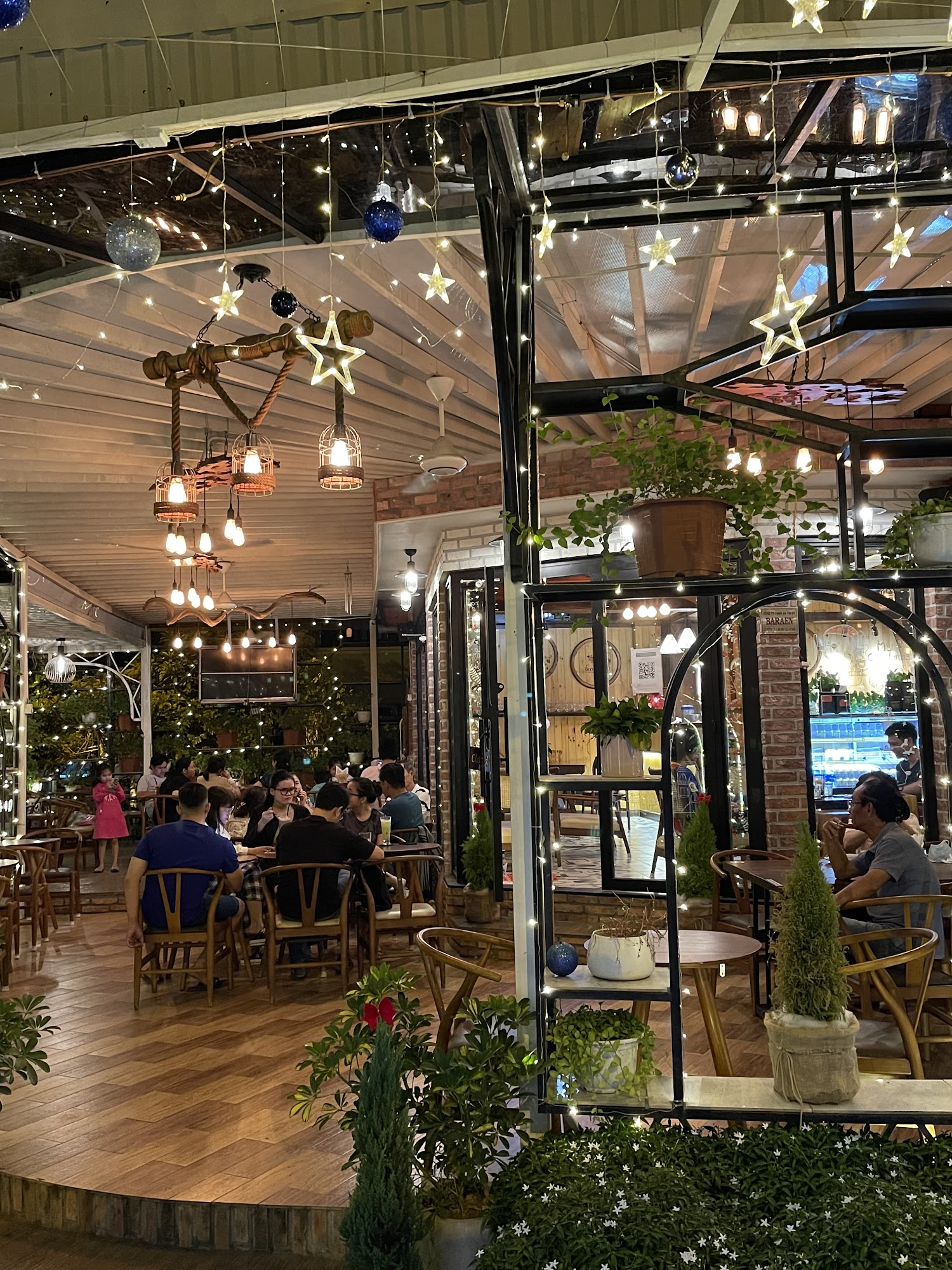 The image depicts the interior of a bustling café or restaurant in Thao Dien, Saigon. It features wooden flooring, exposed brick walls, and a blend of