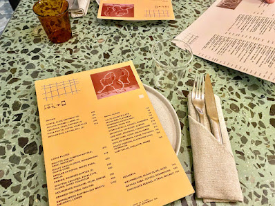 A menu is displayed on a green speckled tablecloth alongside cutlery and a beverage in the foreground.