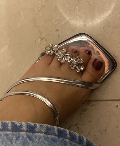 A close-up image of a person's foot wearing silver sandals adorned with floral decorations on the toes and rings around the ankle.