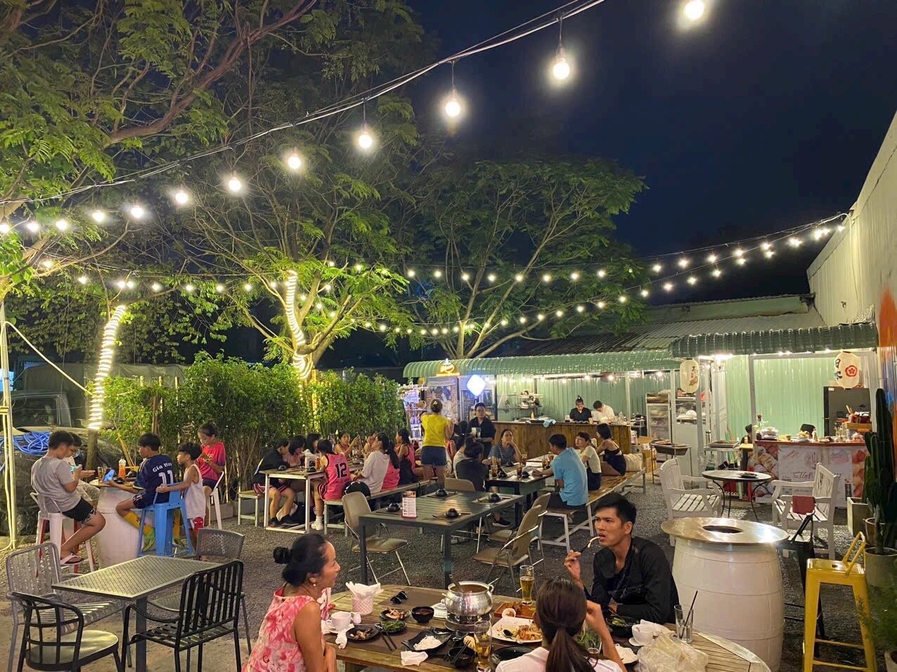 The image depicts an outdoor dining area at night in Thao Dien, Saigon. It is bustling with patrons enjoying their meals under a canopy of string ligh