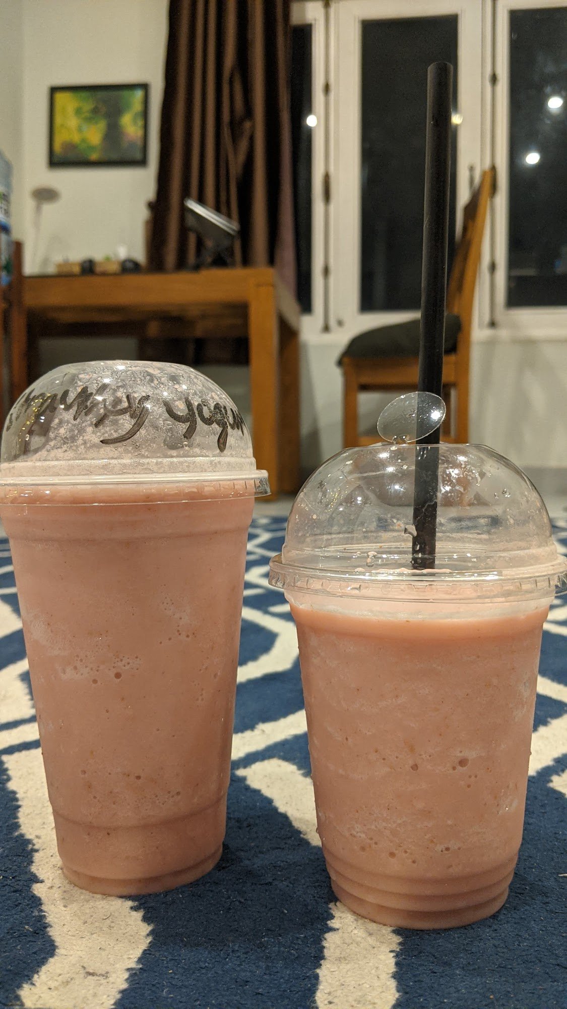 Two beverages in plastic cups on a table; one has a straw and is labeled 'Girly Yogurt'. The setting appears to be indoors, possibly at home.