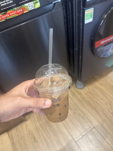 A hand is holding a plastic cup containing an iced coffee beverage with visible ice cubes and dark syrup or flavoring at the bottom, complete with a s