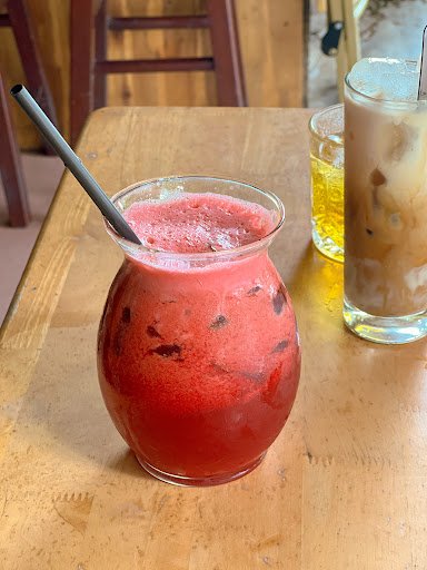 A close-up of a red-colored beverage in a glass jar on a wooden table, with a metal straw and ice cubes visible inside the drink.