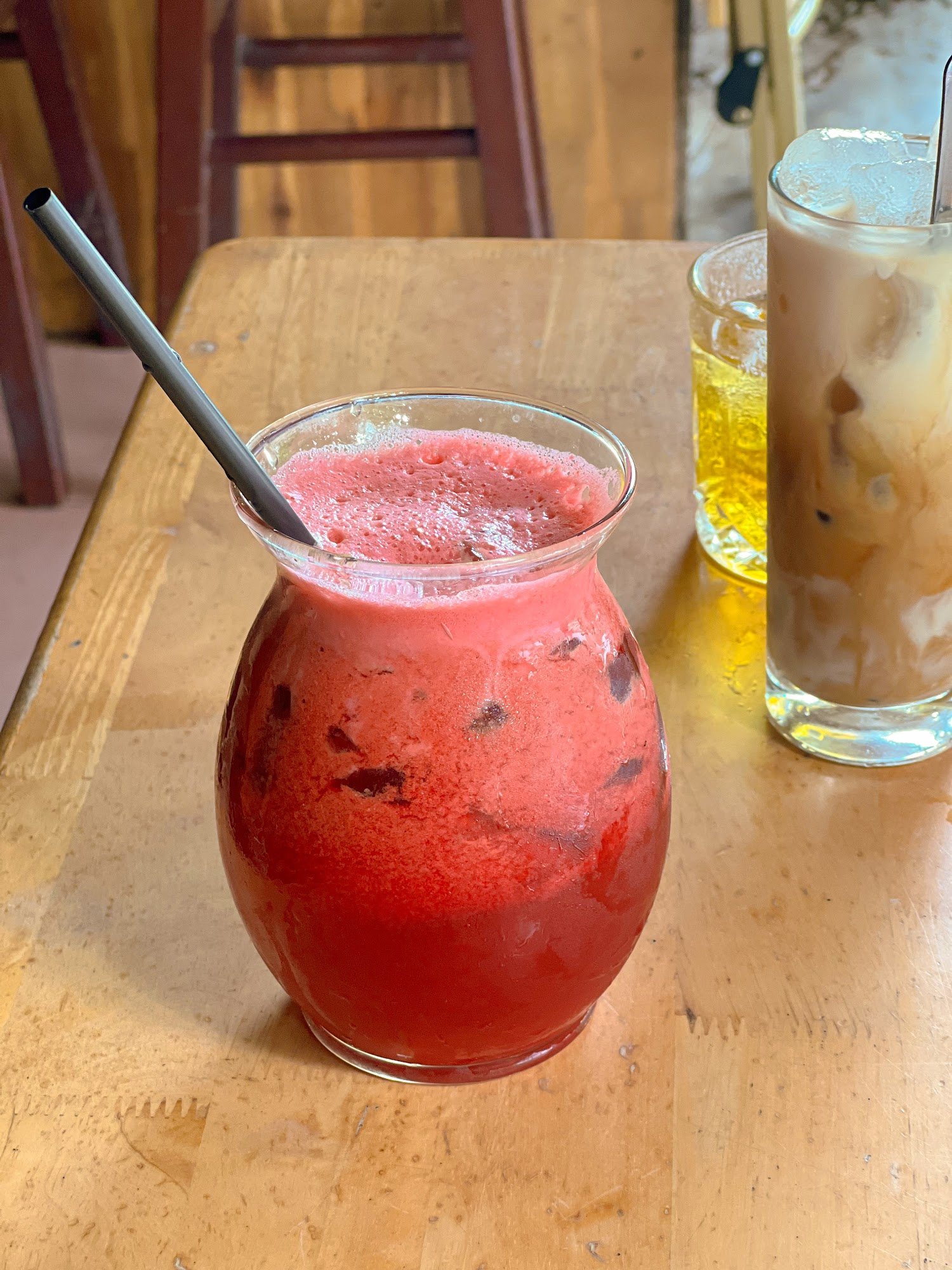 A close-up of a red-colored beverage in a glass jar on a wooden table, with a black straw and ice cubes visible inside the drink.