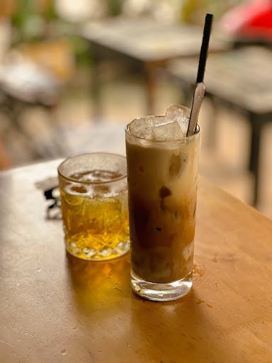 A glass of cold coffee beverage topped with ice cubes and a straw sits on a wooden table next to another drink in a smaller cup, possibly tea or beer.