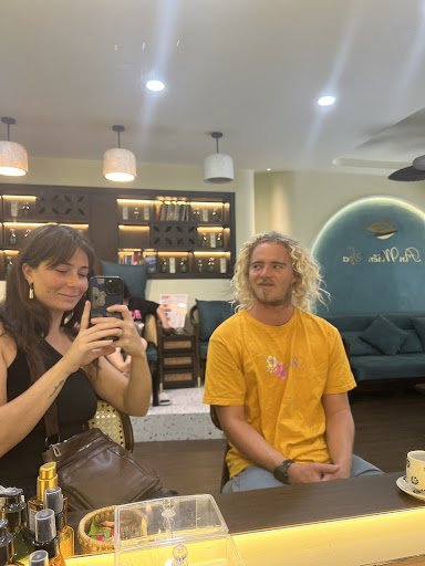 A man and a woman are sitting at the counter of an indoor cafe or coffee shop. The decor includes shelves with bottles, pendant lights on the ceiling,