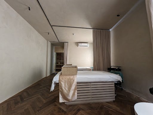 The image shows a modern interior of what appears to be a hotel room or spa suite with wooden flooring and neutral-colored walls. A large bed is posit