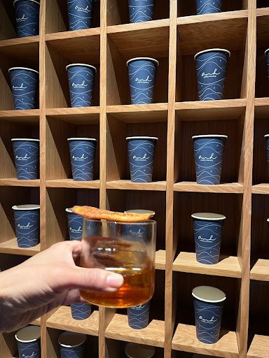 A hand is holding a glass containing an amber-colored liquid and what appears to be a piece of toast or cracker, with multiple blue cups on wooden she