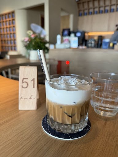 A glass of cold coffee beverage on a wooden table in an indoor setting that appears to be a cafe or restaurant with modern decor, including shelves an