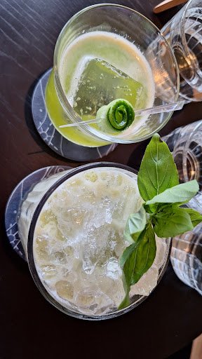 Two glasses of beverages on a dark surface; the top glass contains a green-colored drink with lime and mint garnish, while the bottom glass has a frot