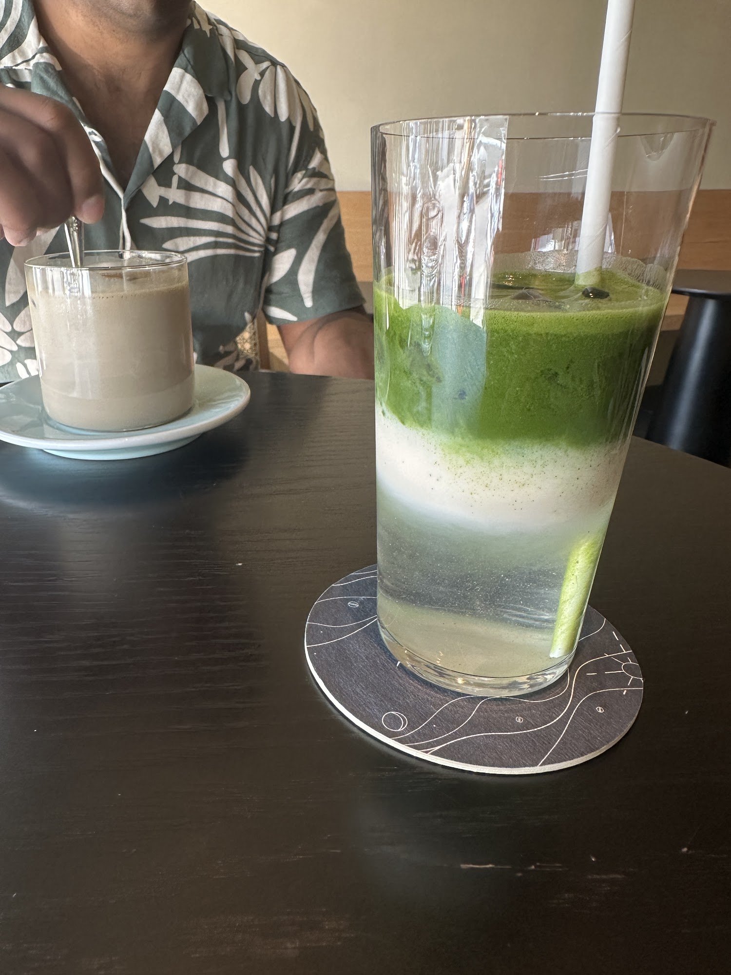 A close-up of a layered green drink in a tall glass on the right and a chocolate milkshake-like beverage with a straw in it to the left, both placed o