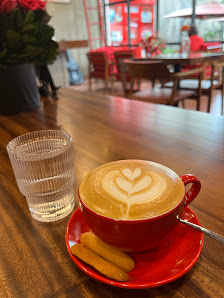 A cozy cafe setting featuring a table with a cup of latte art on a red saucer and spoon, accompanied by two biscotti sticks on the side. A glass of wa