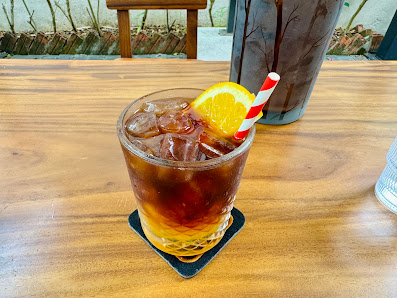 A glass of cola beverage garnished with a slice of orange and a striped straw is placed on a coaster atop a wooden table.