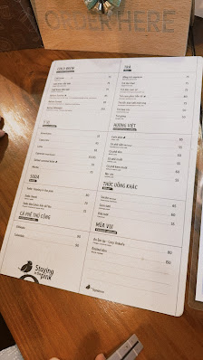 A printed menu from a venue in Thao Dien, Saigon, placed on a wooden table. The menu features sections like 'MAIN DISHES', 'SALADS', 'DRINKS', 'DESSER