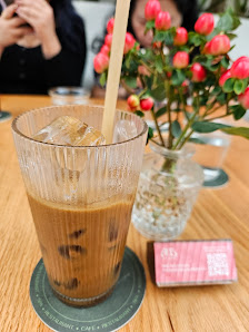 A glass of iced coffee on a table in what appears to be an indoor setting with flowers and greenery visible in the background, suggesting it could be 