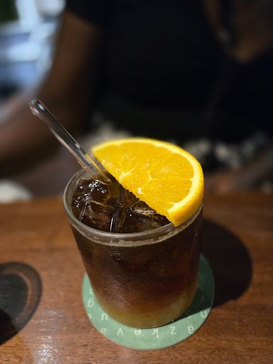 A close-up of a glass containing an amber-colored beverage, possibly cola or tea, with ice cubes and garnished with a slice of orange on top. A clear 
