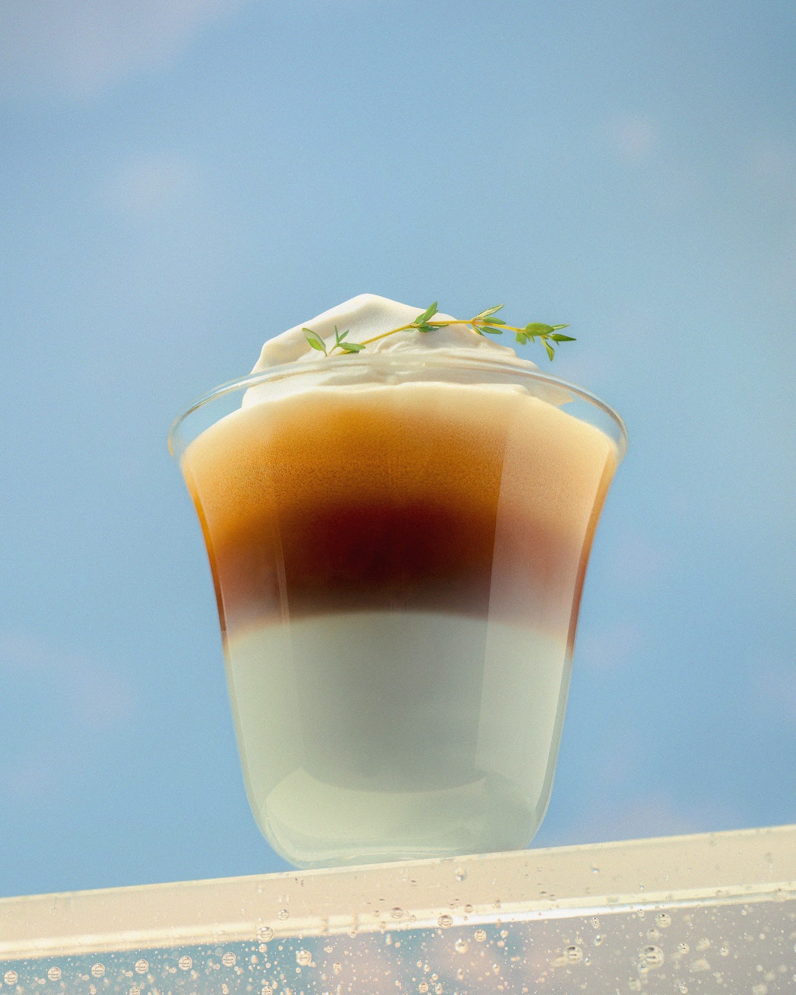 A layered drink in a clear glass topped with whipped cream and garnished with green herbs against a blue sky backdrop.
