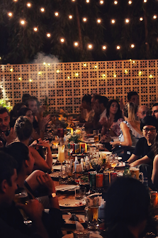A group of people is gathered around a long table outdoors at night for what appears to be a social event or celebration. The setting includes fairy l
