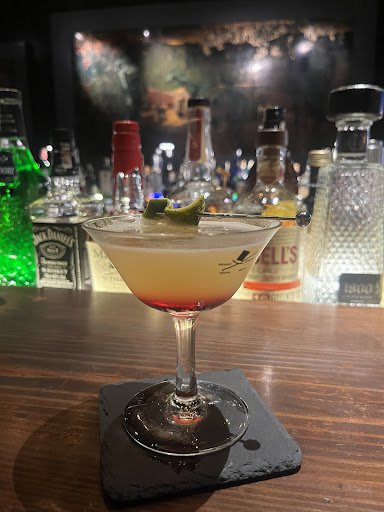 A close-up of a cocktail served in a martini glass on a dark coaster at a bar with various bottles and glasses blurred out in the background.