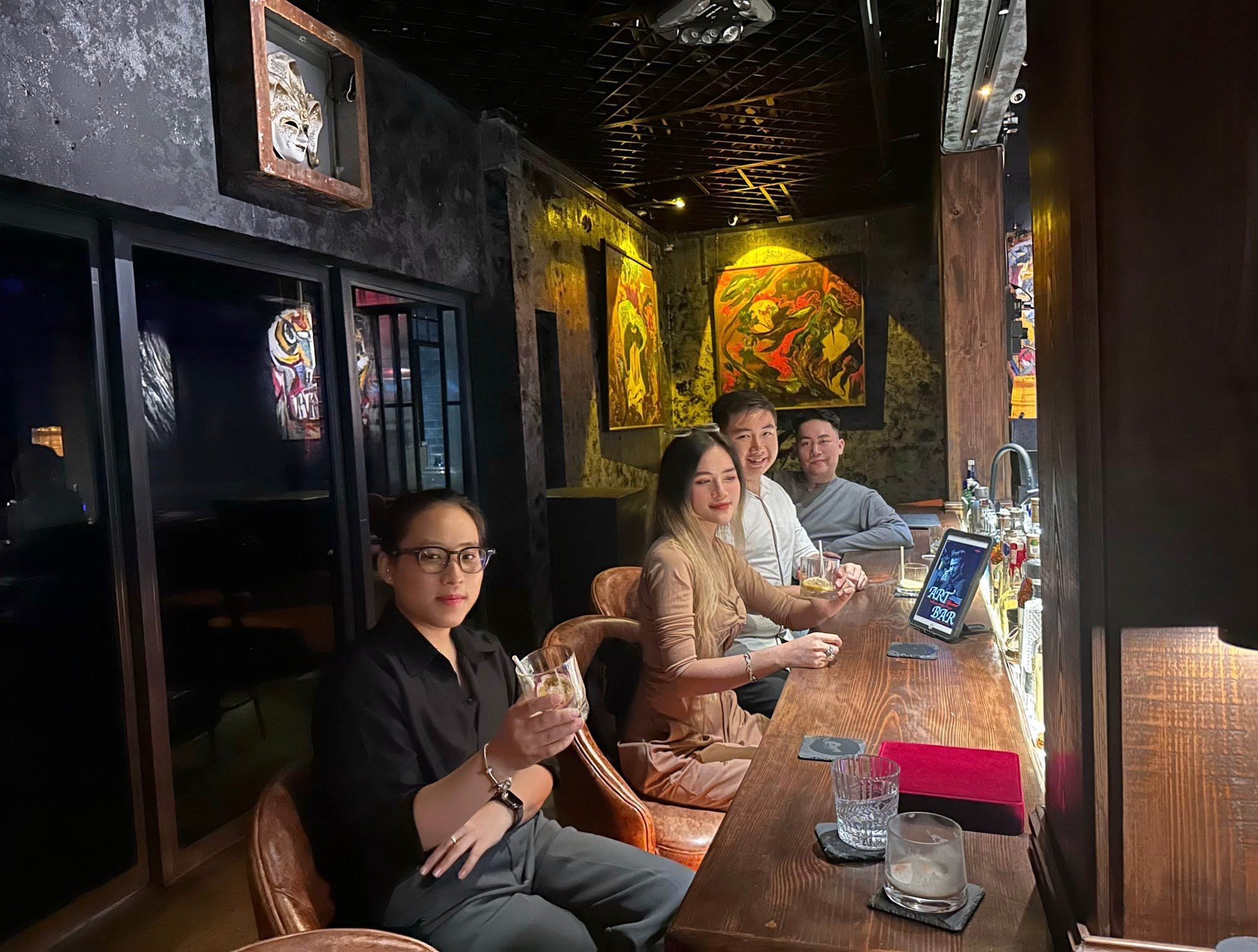 The image shows a group of people sitting at a bar in an indoor venue decorated with vibrant artwork and dim lighting.