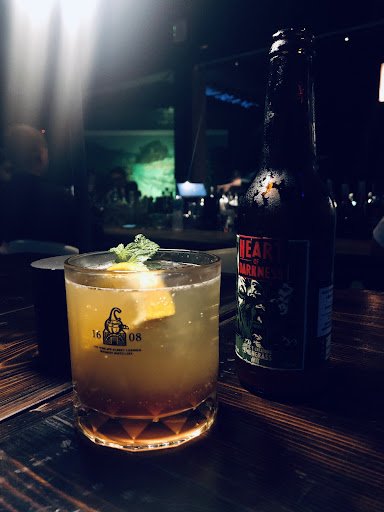 A close-up of a cocktail served in a glass at what appears to be an upscale bar or restaurant setting with dim lighting and ambient background noise, 