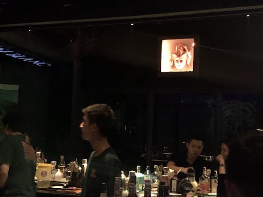 The image captures an indoor scene at a bar or nightclub setting in Thao Dien, Saigon. The focus is on the patrons and the well-stocked bar counter wi