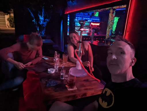 A group of people are sitting at a table in what appears to be a dimly lit bar or lounge with neon lights and colorful decor, engaging in conversation