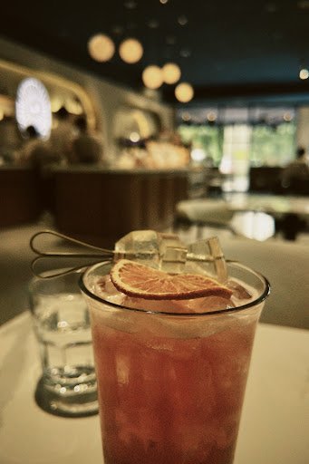 A close-up shot of a refreshing pink cocktail garnished with an orange slice and ice cubes on top, served in a glass at what appears to be a sophistic